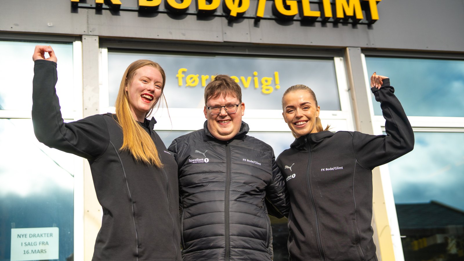 Bildet viser Aubrei Corder og Andrea Holmstrøm fra Bodø/Glimt Kvinner med John-Håkon Nybak som jobber i Glimt-shop i midten, utenfor Glimts supporterbutikk på Aspmyra stadion. Alle smiler og jentene løfter hendene i været i en jubelposisjon.