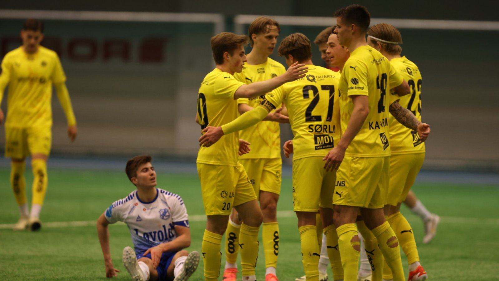 Mange gule Glimt-spillere står sammen og holder rundt hverandre etter at Bodø/Glimt har scoret mot Junkeren. I bakgrunnen sitter en Junkeren-spiller.