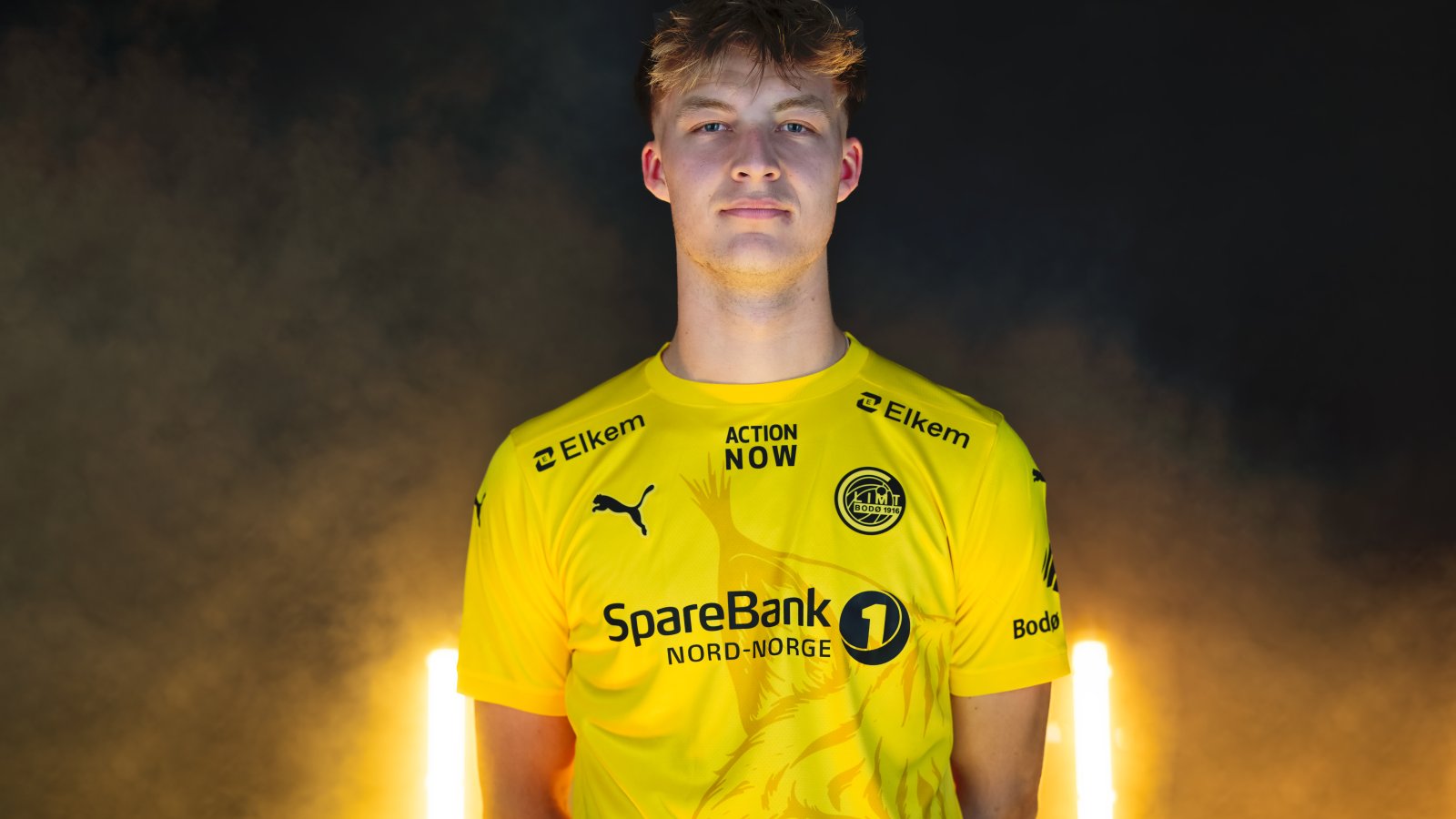 Villads Nielsen poserer i Glimt-drakt etter at han signerte for Bodø/Glimt. I bakgrunnen ser vi gule lys og røyk.